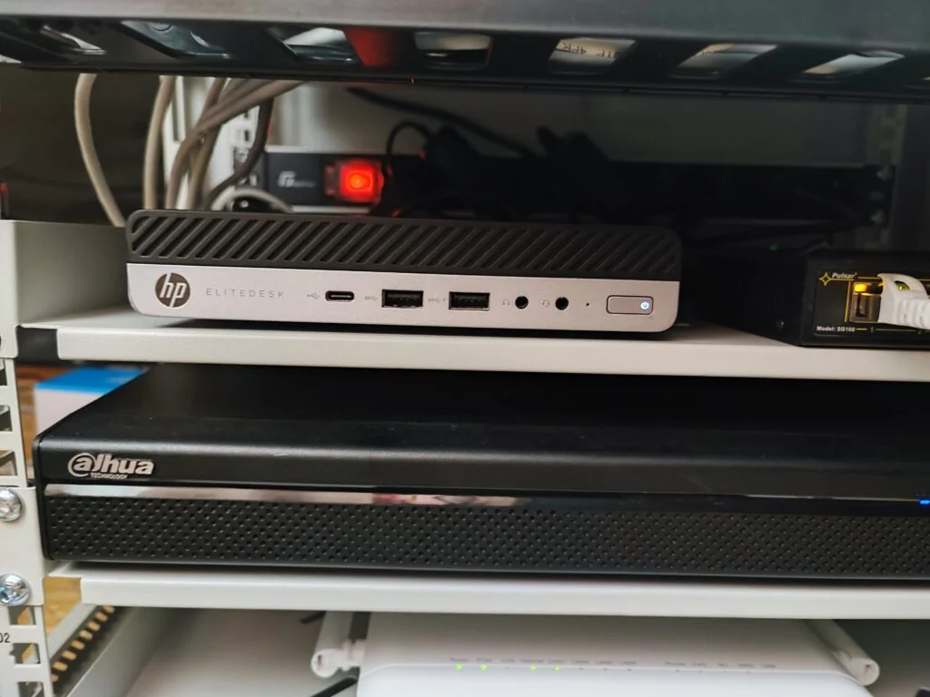 Zbliżenie na komputer HP EliteDesk umieszczony na półce w szafie serwerowej, obok rejestratora Dahua i urządzenia sieciowego, przedstawiające elementy infrastruktury IT.